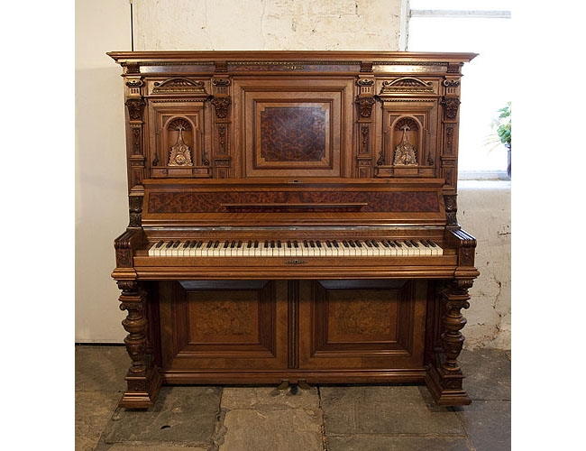 A German upright piano with a Neoclassical style walnut case and cup and cover legs. Cabinet features ornately carved pilasters in high relief and copper sconces in a sea monster design.