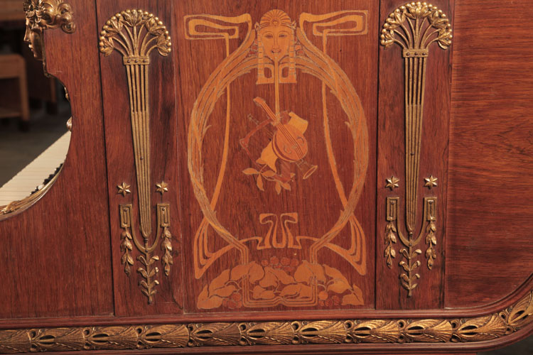The piano cheek side panel is inlaid with an Art Nouveau design featuring a stylised female head surrounded by whiplash lines and foliage. Musical instruments sit centrally in the design.  Columnar, Empire style gilt mounts stand each side of the inlay. Napoleonic emblems of stars, palmettes and foliage feature here
