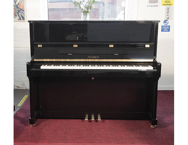 A brand new, Feurich Model 122 upright piano with a black case and chrome fittings. Piano has an eighty-eight note keyboard and three pedals.
