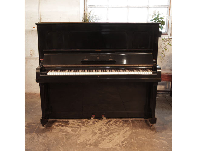 A 1938, Steinway Model K upright piano with a black case and indented front panels. Piano has an eighty-eight note keyboard and two pedals.  